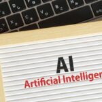 Wooden-framed sign reading 'AI' and 'Artificial Intelligence' on a striped background with a laptop keyboard in the background.