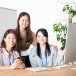 Three coworkers smile at the camera in a bright office, gathered around a desk with a computer and a plant in the background.