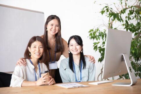 Three coworkers smile at the camera in a bright office, gathered around a desk with a computer and a plant in the background.