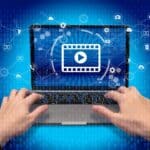 Hands typing on a laptop with a video play icon on the screen, surrounded by digital icons.