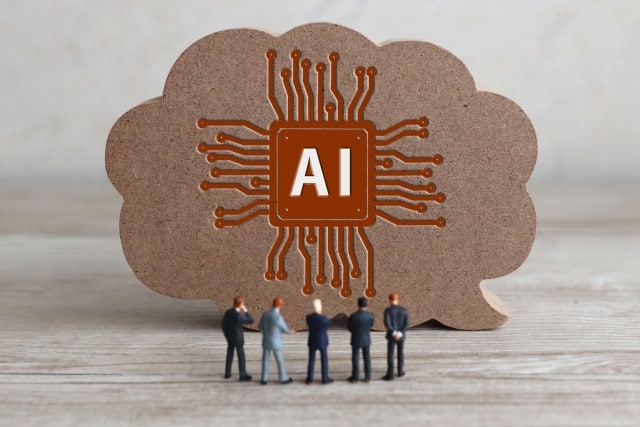 Cloud-shaped cardboard with an orange AI circuit logo and miniature people gathered in front, signaling AI teamwork.