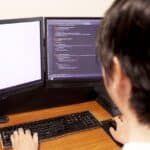 Person coding at a dual-monitor workstation, left screen blank and right screen showing code, with keyboard and headphones on a wooden desk.