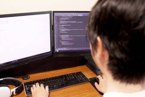 Person coding at a dual-monitor workstation, left screen blank and right screen showing code, with keyboard and headphones on a wooden desk.