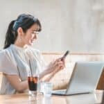 Woman at a cafe table using her phone with a laptop open in front of her and a glass of iced coffee nearby.
