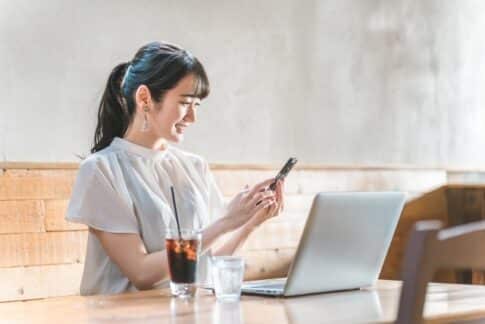Woman at a cafe table using her phone with a laptop open in front of her and a glass of iced coffee nearby.