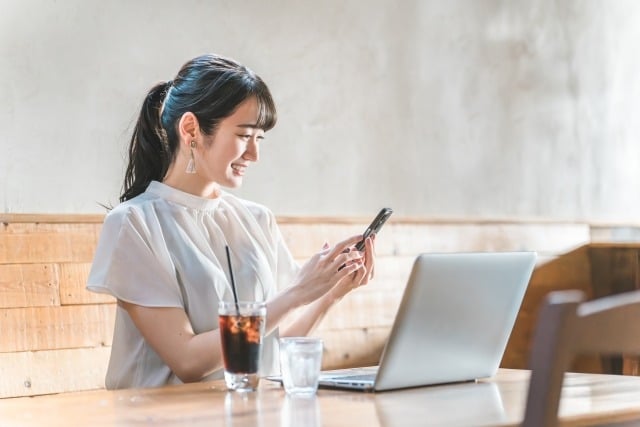 Woman at a cafe table using her phone with a laptop open in front of her and a glass of iced coffee nearby.