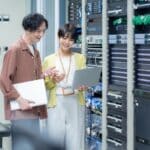 Two IT professionals review server rack hardware with a laptop in a data center, collaborating.