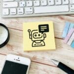 Sticky note with a simple robot illustration and speech bubble on a wooden desk, near a keyboard, smartphone, pen, magnifying glass, and color swatches.