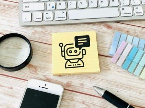 Sticky note with a simple robot illustration and speech bubble on a wooden desk, near a keyboard, smartphone, pen, magnifying glass, and color swatches.