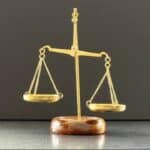 Gold balance scale on a wooden base, symbolizing justice or fairness, with weights balanced evenly against a dark backdrop.