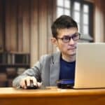 Man in glasses and a blazer uses a laptop and mouse at a wooden desk in a cozy office setting, looking focused.