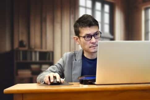 Man in glasses and a blazer uses a laptop and mouse at a wooden desk in a cozy office setting, looking focused.