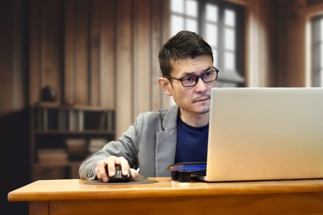 Man in glasses and a blazer uses a laptop and mouse at a wooden desk in a cozy office setting, looking focused.