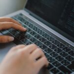Close-up of hands typing on a laptop keyboard with code visible on the screen.