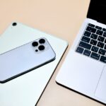 Triple-camera iPhone resting on a light-green laptop, with another silver laptop keyboard shown nearby on a beige desk