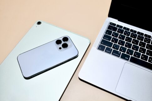 Triple-camera iPhone resting on a light-green laptop, with another silver laptop keyboard shown nearby on a beige desk