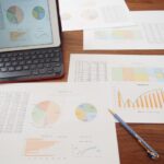 Tablet displaying color charts beside printed reports with pie, bar, and line graphs on a wooden desk.