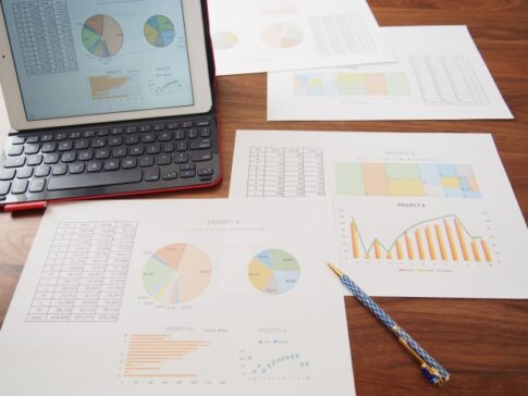 Tablet displaying color charts beside printed reports with pie, bar, and line graphs on a wooden desk.