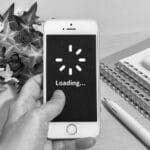 Hand holding an iPhone with a loading spinner and 'Loading...' on screen, on a desk with notebooks, pen, calculator, and a plant nearby