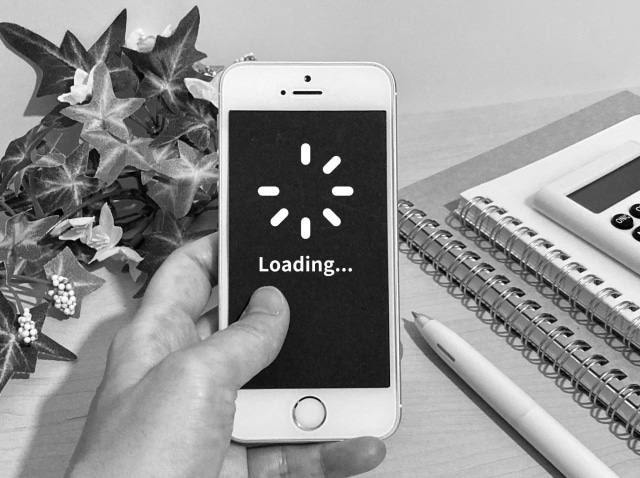 Hand holding an iPhone with a loading spinner and 'Loading...' on screen, on a desk with notebooks, pen, calculator, and a plant nearby